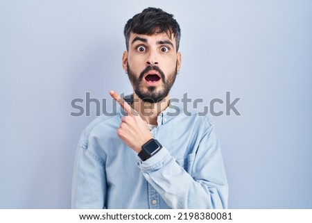 Young hispanic man with beard standing over blue background surprised pointing with finger to the side, open mouth amazed expression. 