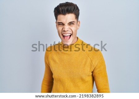 Young hispanic man standing over blue background angry and mad screaming frustrated and furious, shouting with anger. rage and aggressive concept. 
