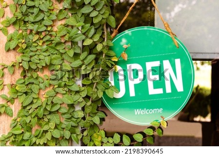 Hanging green sign with message OPEN on window with leaves ivy on wooden wall at coffee shop