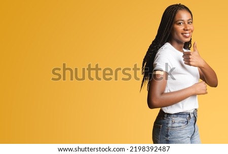 Happy young african-american woman with medical patch aid on the forearm showing thumbs up in sign approving of vaccination, recomending keep self in safety from viral disease