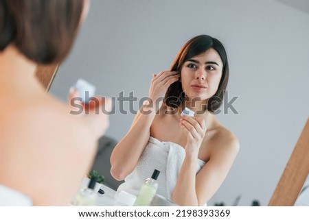 Sitting by the mirror and using cream on face. Young woman is at home. Conception of beauty.