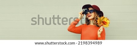 Autumn portrait of happy smiling woman with film camera and yellow maple leaves wearing red sweater and hat on gray background, blank copy space for advertising text