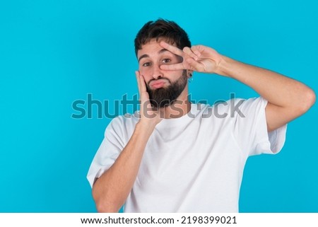 bearded caucasian man wearing white T-shirt over blue background  making v-sign near eyes. Leisure lifestyle people person celebrate flirt coquettish concept.