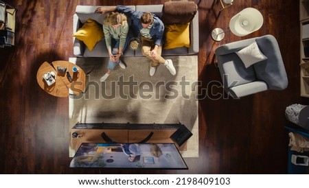 Top View Apartment: Happy Couple Watching Television in the Stylish Living Room. Looking at the TV Display and Eating Popcorn. Girlfriend and Boyfriend Enjoy Sitcom on Streaming Service on Couch. Royalty-Free Stock Photo #2198409103