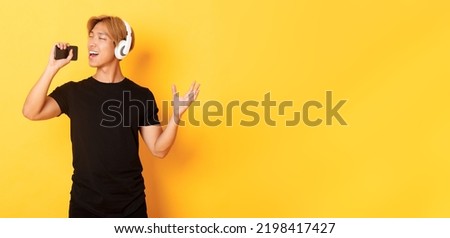Joyful attractive korean guy in headphones, playing karaoke app, singing into mobile phone microphone, standing yellow background