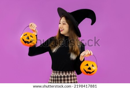Halloween, young asian girl in black costume wearing black witch hat holding and carrying orange pumpkin bucket on purple background.