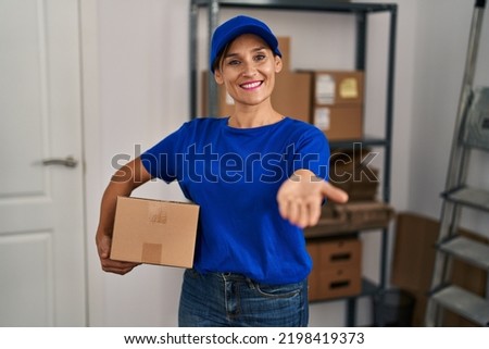 Middle age brunette woman working wearing delivery uniform and cap smiling cheerful offering palm hand giving assistance and acceptance. 