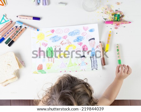 Little girl choosing a green pencil in a wood table. Multi-racial Family drawing on a white paper witch coloured pencils, a sharpener, chalks and a sandwich that has been bitten once