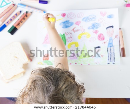 Little girl painting a sun in a kids drawing Family. Multi-racial Family drawing on a white paper witch coloured pencils, a sharpener and a sandwich that has been bitten once