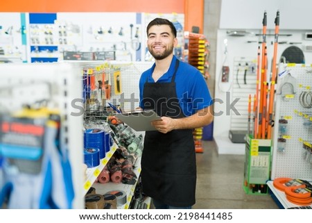 Cheerful retail worker with a blue uniform smiling holding a clipboard for inventory and looking happy to work at the hardware store  Royalty-Free Stock Photo #2198441485
