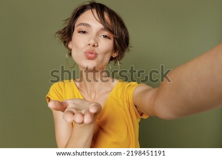 Close up young fun woman she 20s wear yellow t-shirt doing selfie shot pov on mobile cell phone blow air kiss isolated on plain olive green khaki background studio portrait. People lifestyle concept