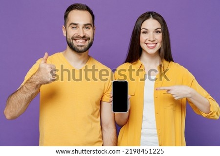 Young couple two friends family man woman together in yellow clothes hold use point index finger on mobile cell phone with blank screen workspace area show thumb up isolated on plain violet background