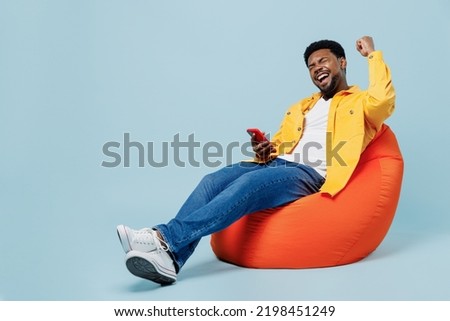 Full body young man of African American ethnicity in yellow shirt sit in bag chair hold in hand use mobile cell phone do winner gesture isolated on plain pastel light blue background studio portrait.