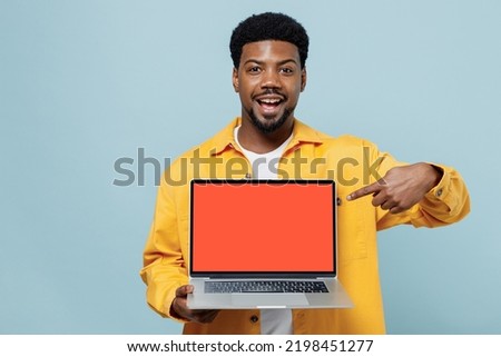 Young happy man of African American ethnicity wear yellow shirt hold use work point index finger on laptop pc computer with blank screen workspace area isolated on plain pastel light blue background