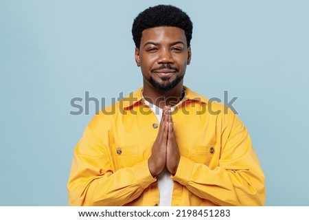 Young man of African American ethnicity 20s in yellow shirt hold hands folded in prayer gesture, begging about something isolated on plain pastel light blue background studio. People lifestyle concept Royalty-Free Stock Photo #2198451283