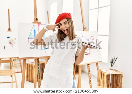 Young artist woman painting on a canvas at art studio doing peace symbol with fingers over face, smiling cheerful showing victory 