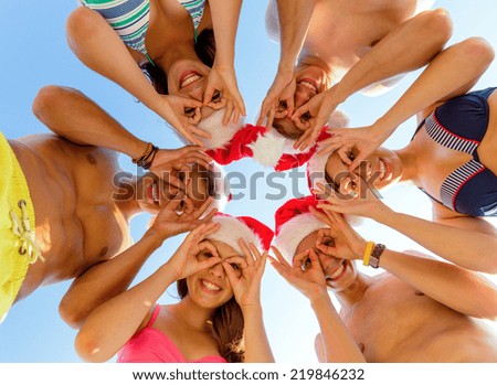 friendship, christmas, summer vacation, holidays and people concept - group of smiling friends wearing swimwear and santa helper hats standing and having fun in circle over blue sky