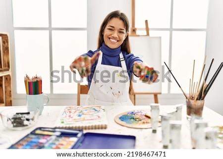 Young brunette woman at art studio with painted hands approving doing positive gesture with hand, thumbs up smiling and happy for success. winner gesture. 
