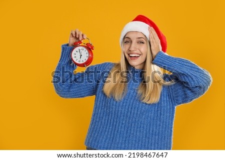 Woman in Santa hat with alarm clock on yellow background. New Year countdown