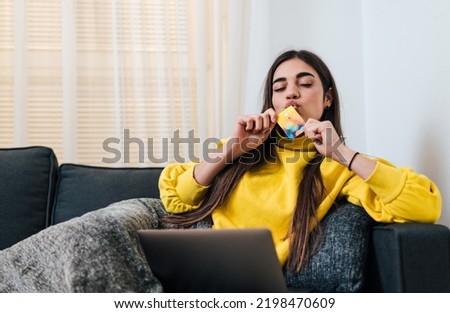 Relaxed young brunette woman, bringing her credit card close to her lips, wearing yellow sweatshirt.