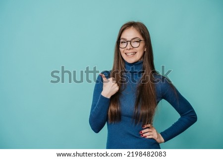 Attractive Italian young caucasian woman in blue sweater and glasses smiling wide, showing thumb up gesture over studio turquoise background with empty space for ad. Mockup, shopping, sales, discount.
