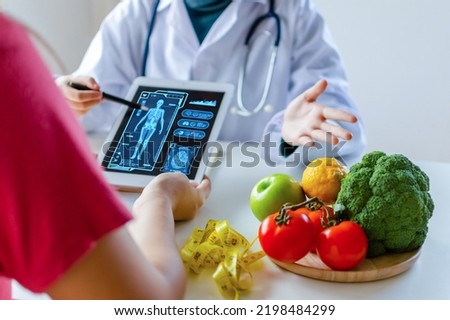 nutritionist female doctor using digital mobile tablet with virtual graphic icon diagram and vegetable and fruit with patient on desk at office hospital, nutrition, food science, healthy food concept Royalty-Free Stock Photo #2198484299