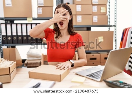 Young hispanic woman preparing order working at storehouse peeking in shock covering face and eyes with hand, looking through fingers with embarrassed expression. 