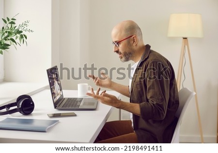 Man having casual online business meeting at home. Side view of man in glasses sitting in front of modern laptop computer in his homeoffice, gesturing and discussing work with colleague on videocall Royalty-Free Stock Photo #2198491411
