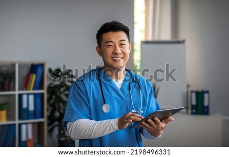 Smiling middle aged japanese male doctor with tablet and wait patient in clinic office interior. Treatment with professional therapist, medical health care and good service during covid-19 pandemic Royalty-Free Stock Photo #2198496331
