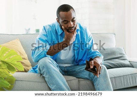 I'm broke. Sad african american man bankrupt showing his empty wallet and looking at it, sitting on sofa at home, free space. Poverty concept Royalty-Free Stock Photo #2198496679