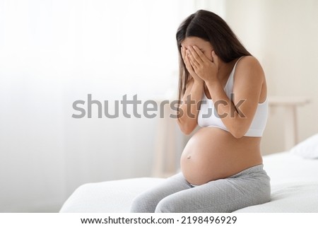Pregnancy hormonal changes concept. Young long-haired pregnant woman in homewear with big tummy sitting alone on bed at home, covering her face, feeling sad and crying, free space Royalty-Free Stock Photo #2198496929