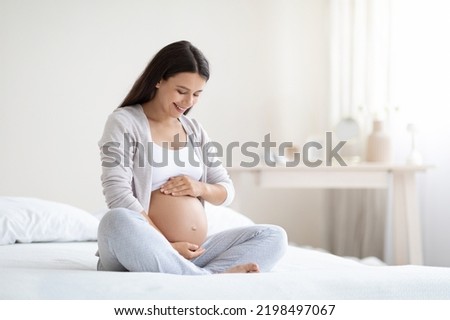 Happy pregnant woman sitting on bed in bedroom at home, hugging her big tummy. Happiness and feeling joyful as emotional well affect to baby neurological and psychological development, empty space Royalty-Free Stock Photo #2198497067