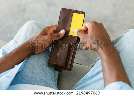 African american man selecting credit card from his card wallet, sitting on couch at home, closeup. Modern payments and online banking concept