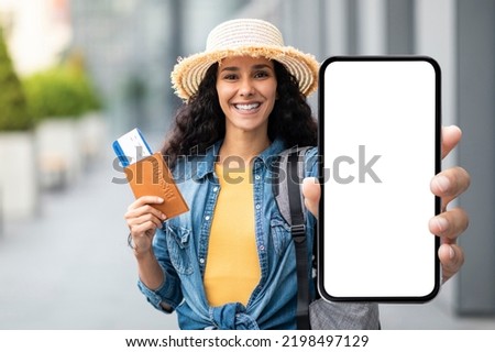 Happy attractive millennial brunette lady in straw hat tourist holding passport with flight tickets and cell phone with white blank screen, showing nice mobile app for travellers, mockup Royalty-Free Stock Photo #2198497129
