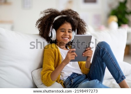Happy african american kid school girl with bushy hair sitting on couch, using digital tablet and wireless headset, playing mobile game or checking newest app, home interior, copy space