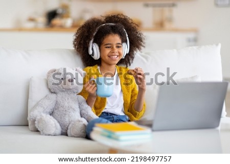 Cute black school girl having break while doing homework, child sitting on couch, drinking cocoa and eating cookies, using wireless headphones, looking at laptop screen and smiling, copy space