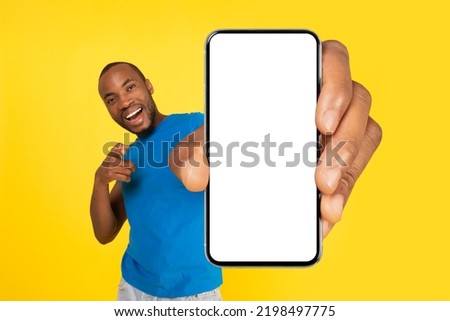 Joyful Black Guy Holding Phone With Large Empty Screen Advertising Mobile Offer Pointing Finger Standing In Studio On Yellow Background. Check This Great Application Concept. Mockup Royalty-Free Stock Photo #2198497775
