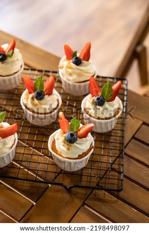 Cupcakes with white icing decorated with strawberries, blueberries and mint on metal grille. Delicious homemade dessert. Festive bakery. Blurred background.