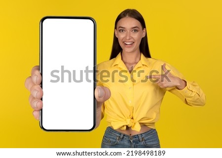 Mobile Offer. Positive Lady Showing Huge Smartphone With Empty Screen Advertising Application, Pointing At Phone Standing Over Yellow Background In Studio. New Application Advert. Mockup