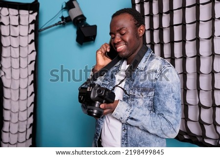 Photographer talking on smartphone while standing on blue background. Young producer having a phone call while working on production studio equipped with professional softboxes and spotlights.
