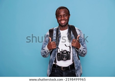 Happy photography enthusiast showing approval finger gesture while having DSLR photo device on blue background. Smiling young man with professional camera giving thumbs up hand symbol.