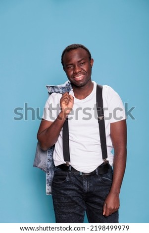 Cheerful young adult man having denim jacket while smiling at camera and standing on blue background. Handsome and confident looking guy posing for camera while wearing suspenders and tshirt.