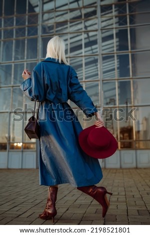 Rear, back view of fashionable blonde woman wearing long blue trench coat, marsala color leather ankle boots, holding stylish hat, bag, walking in street of city. Female fashion conception Royalty-Free Stock Photo #2198521801