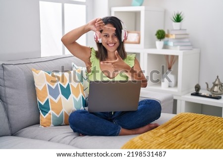 Hispanic young woman using laptop at home smiling making frame with hands and fingers with happy face. creativity and photography concept. 