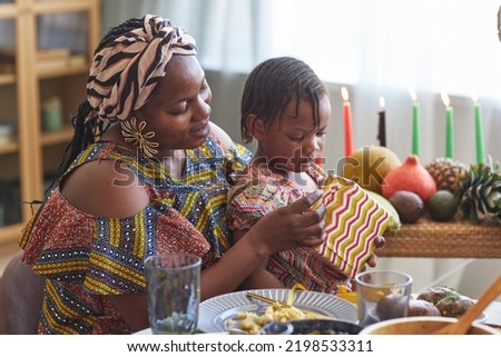 African woman in national costume opening present with her little daughter while they sitting at dining table