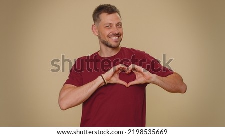 I love you. Smiling bearded handsome young man makes heart gesture demonstrates love sign expresses good feelings and sympathy. Adult stylish male guy isolated on beige studio background indoors