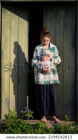 Young girl in the countryside in summer. White-red cat is in her hands. In the background are large green wooden doors. Girl stands barefeet. Beautiful young woman outdoors. Vacation in the village.