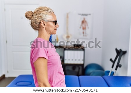 Middle age blonde woman at pain recovery clinic looking to side, relax profile pose with natural face with confident smile. 