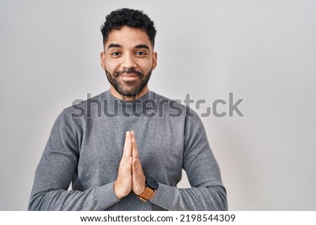 Hispanic man with beard standing over white background praying with hands together asking for forgiveness smiling confident. 