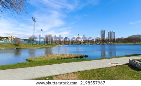 There are trees and grass on the shore of the lake. Along the shore is a paved sidewalk. On the opposite shore is a stadium with lighting poles, an Orthodox church and apartment buildings. Sunny Royalty-Free Stock Photo #2198557967